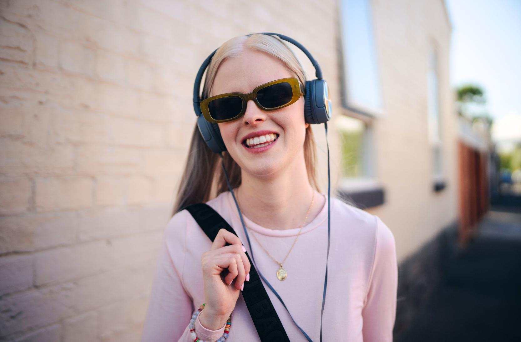 A blonde woman wearing a pink top, headphones and sunglasses smiles to camera.